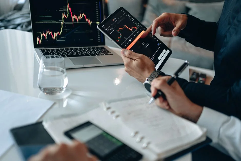 Hands using smartphone beside laptop with stock charts, showcasing digital trading.